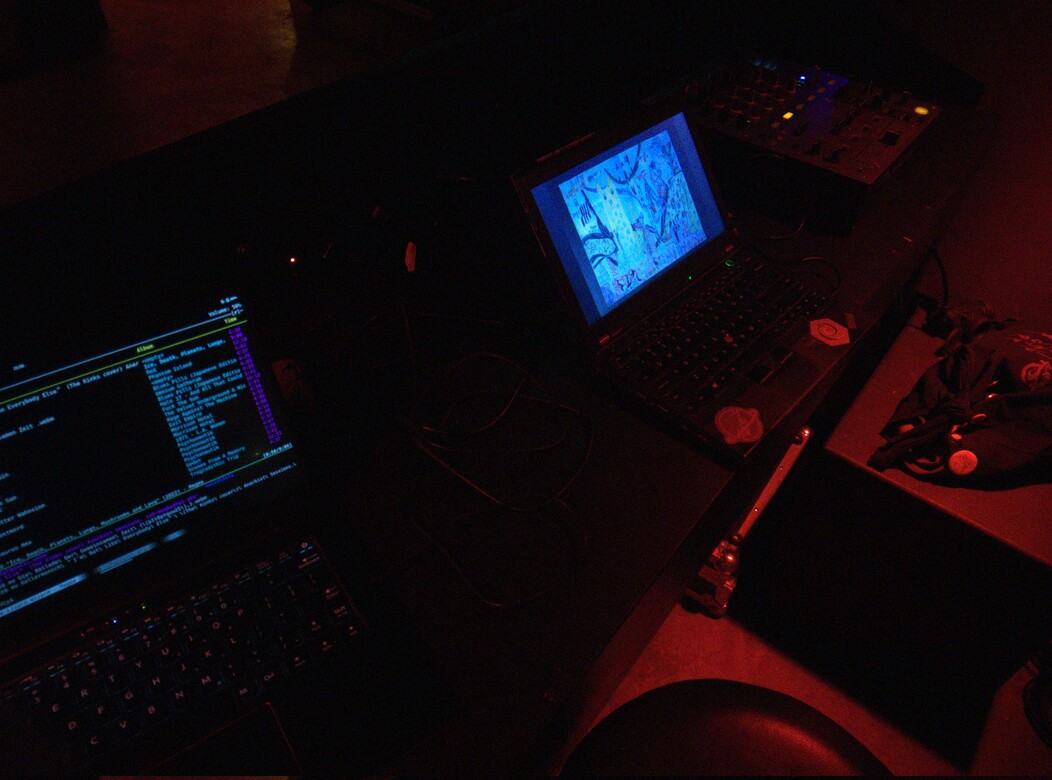 Two laptops on a black table connected to a audio mixer in a corner that is lit dimly red Laptops providing music to the sound system