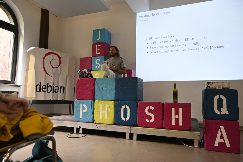 Manuel on stage. His slides areprojected against the wall behind him. The stage decoration includes a large banner with the Debian swirl and some cubes are arranged to spell the words 'PHOSH' and 'QA' respectively. Manuel presenting his talk on immutable images